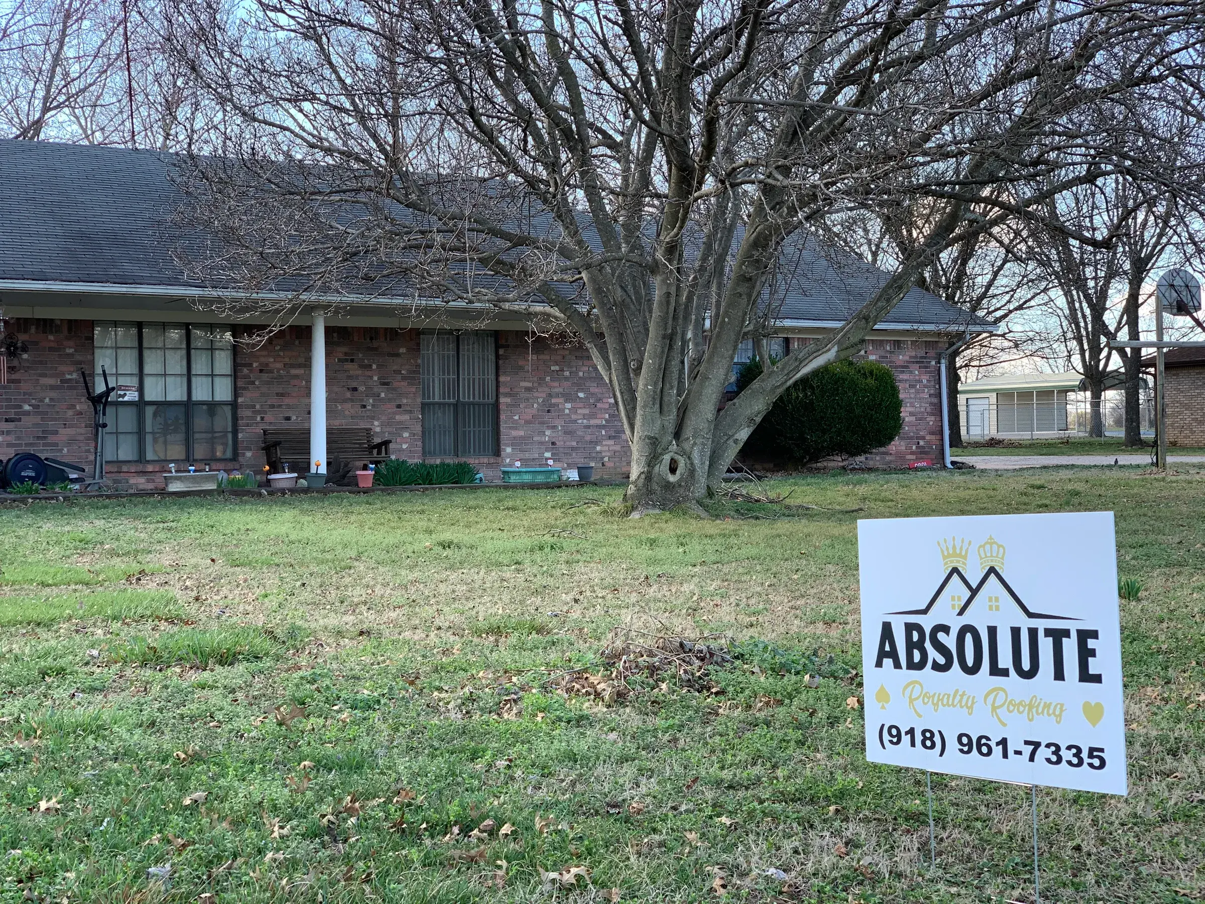 An Absolute Royalty Roofing yard sign in the front lawn of a brick ranch house with a dark shingle roof, photographed on a bare winter day.