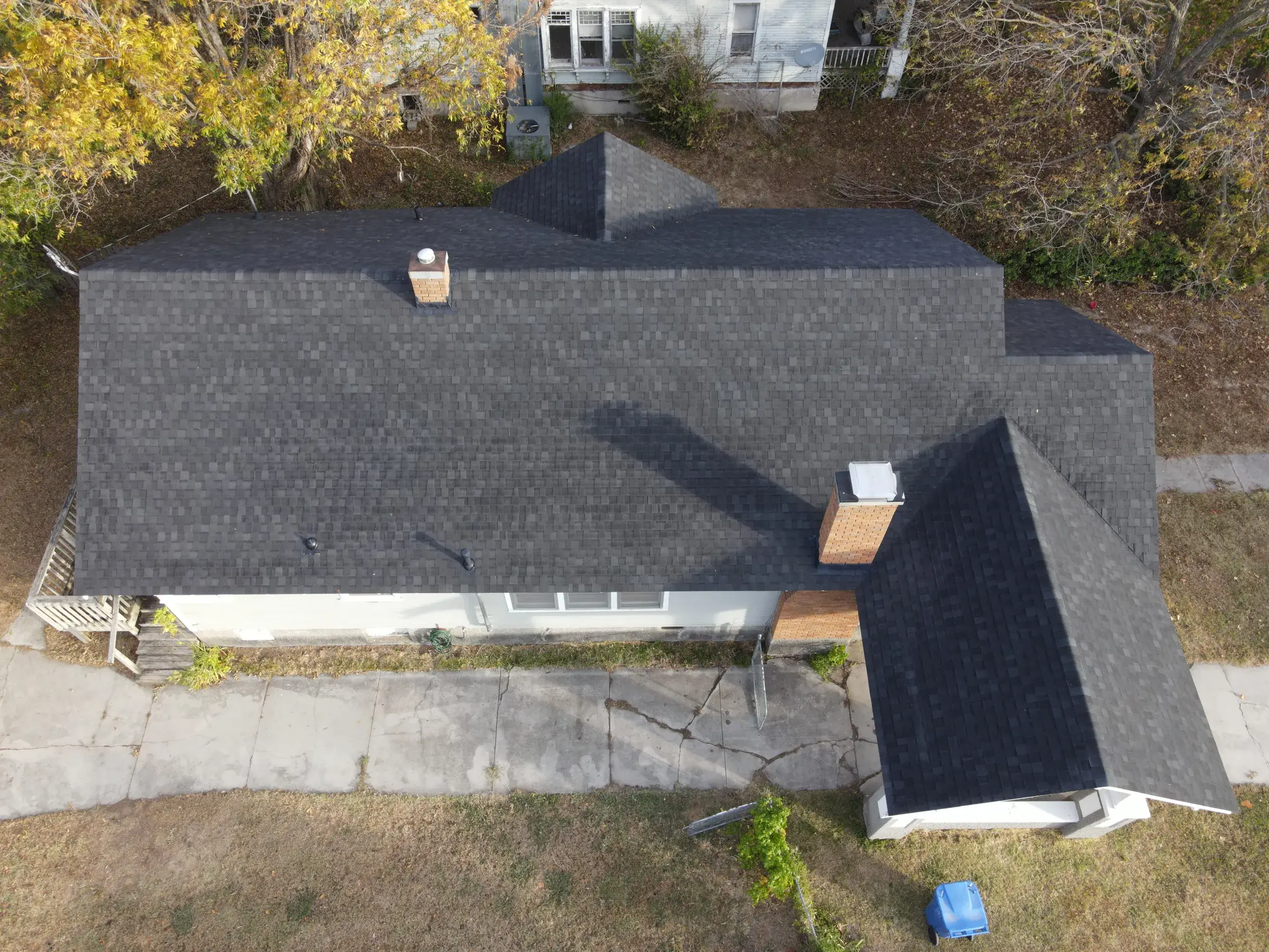 Overhead drone view of a single-story home with a dark charcoal asphalt shingle roof, a brick chimney, and a small wooden deck, surrounded by autumn trees.