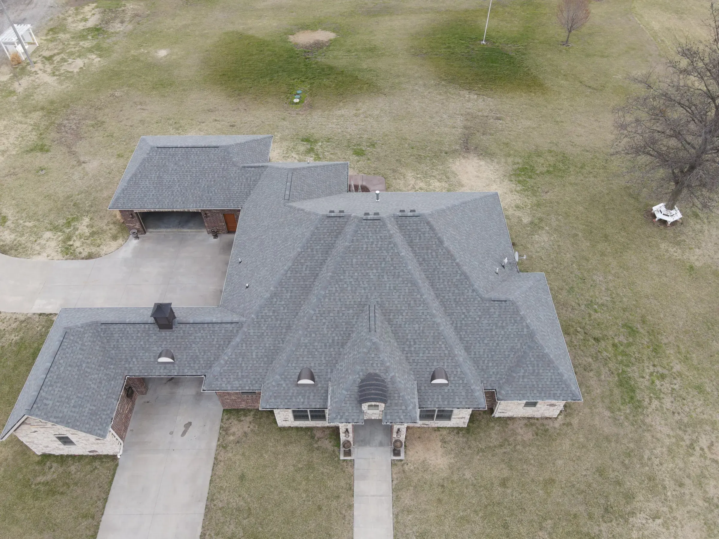Aerial view of a large brick ranch home on rural acreage with a gray architectural asphalt shingle roof and wide concrete driveways.