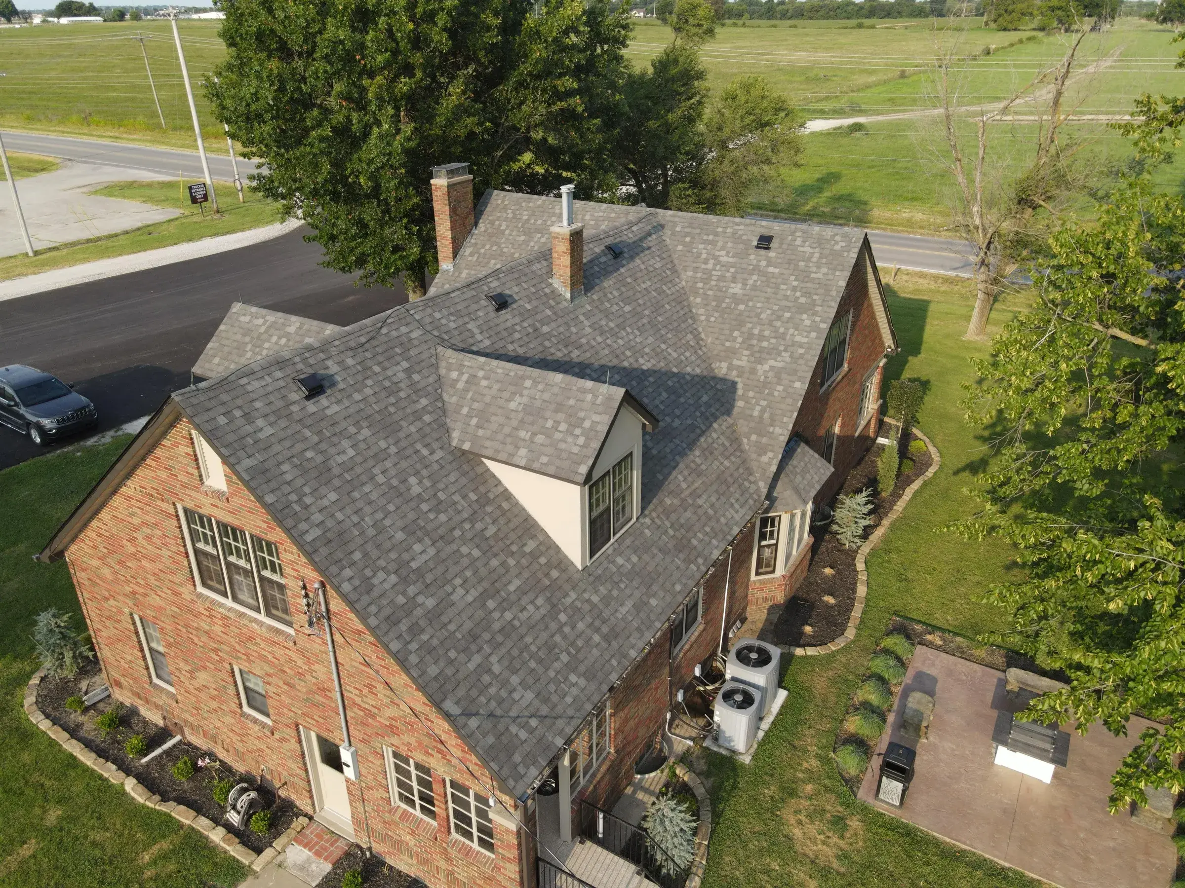 Aerial view of a two-story red brick house with a newly installed gray shingle roof, surrounded by mature trees and open green fields.