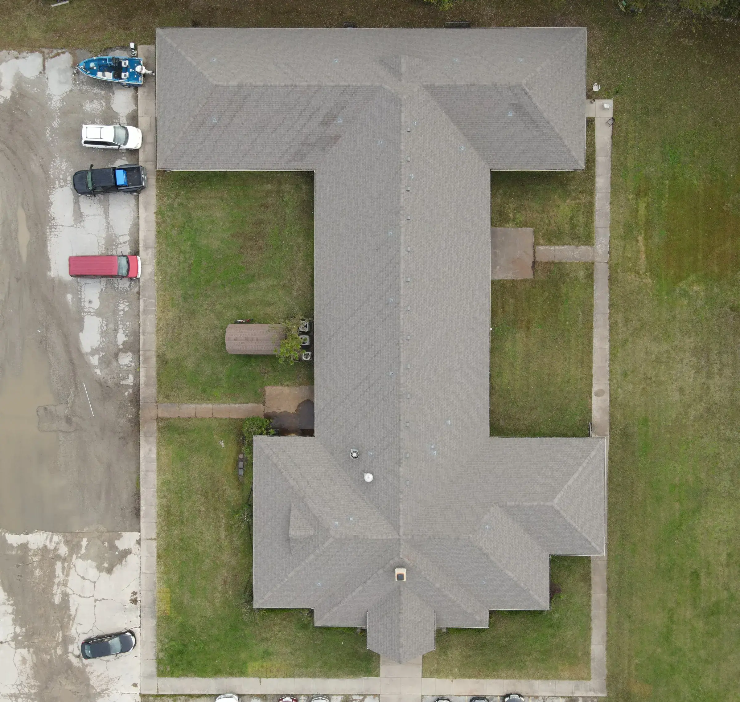 Overhead drone view of a large H-shaped house with a gray shingle roof, with vehicles and a boat parked on the gravel drive alongside it.
