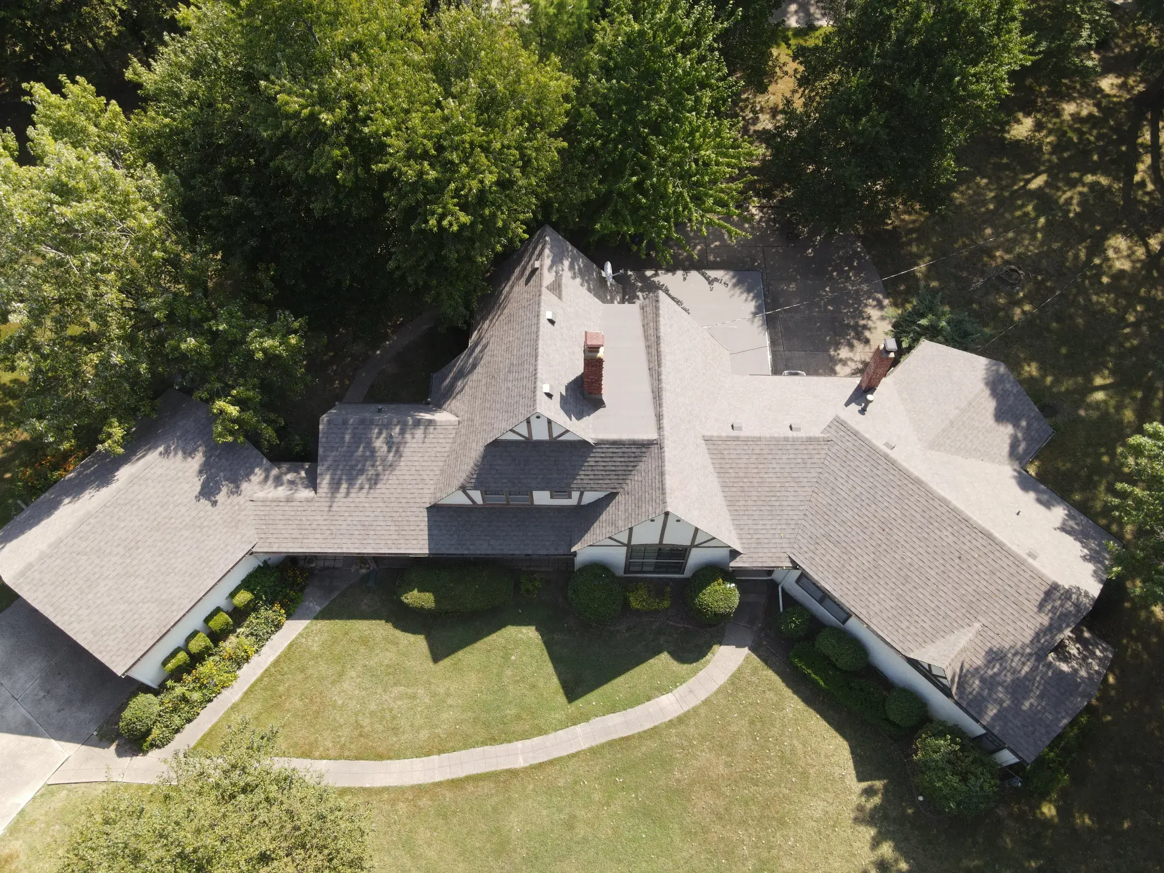 Aerial view of a large single-story home with a sprawling light gray shingle roof, two brick chimneys, and a glass sunroom, surrounded by manicured lawn and mature trees.