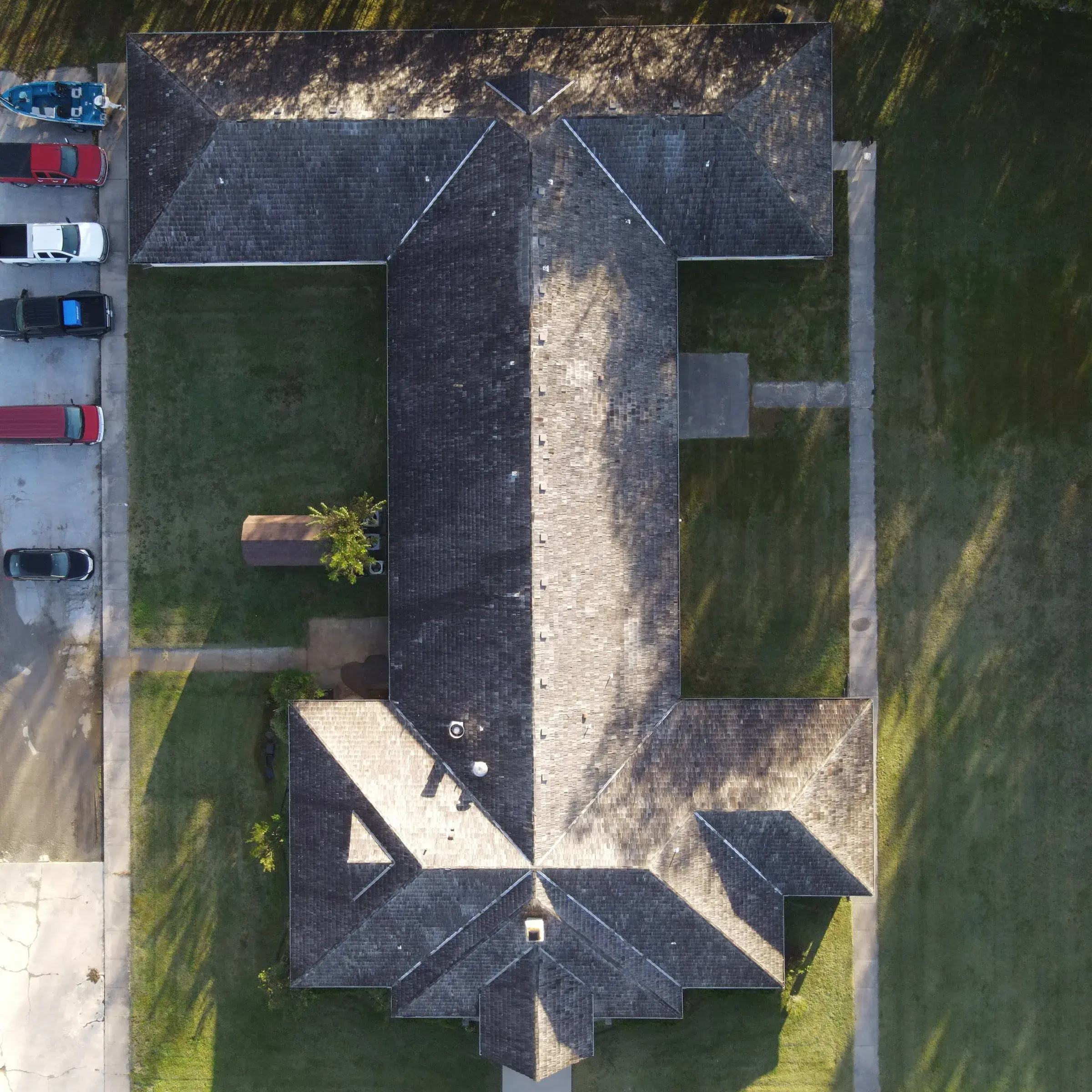 Overhead drone view of a large home's dark, weathered asphalt shingle roof with multiple hips and valleys, with vehicles parked along the drive at left.