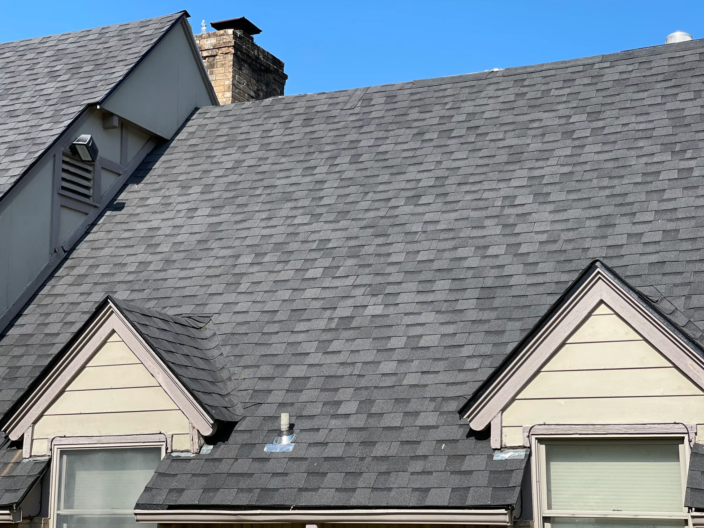 Close-up of a newly laid charcoal architectural shingle roof with two dormers and a brick chimney against a clear blue sky.