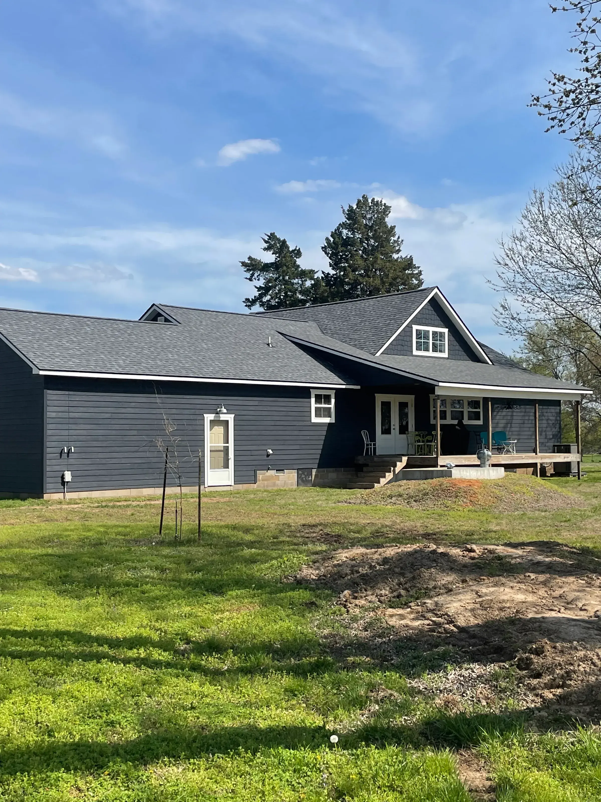 Rear view of a dark navy farmhouse-style home with a dark shingle roof and covered porch, with freshly turned soil in the yard.