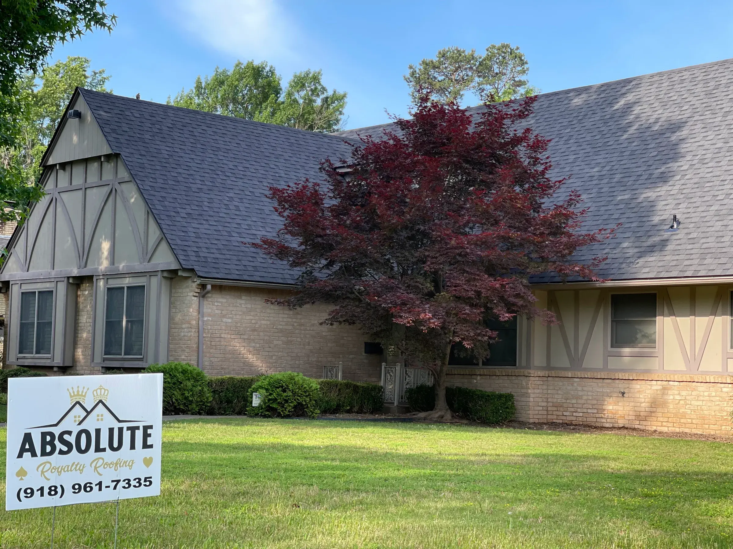 A Tudor-style brick home with a dark charcoal asphalt shingle roof and a red Japanese maple in front, with an Absolute Royalty Roofing yard sign in the lawn.