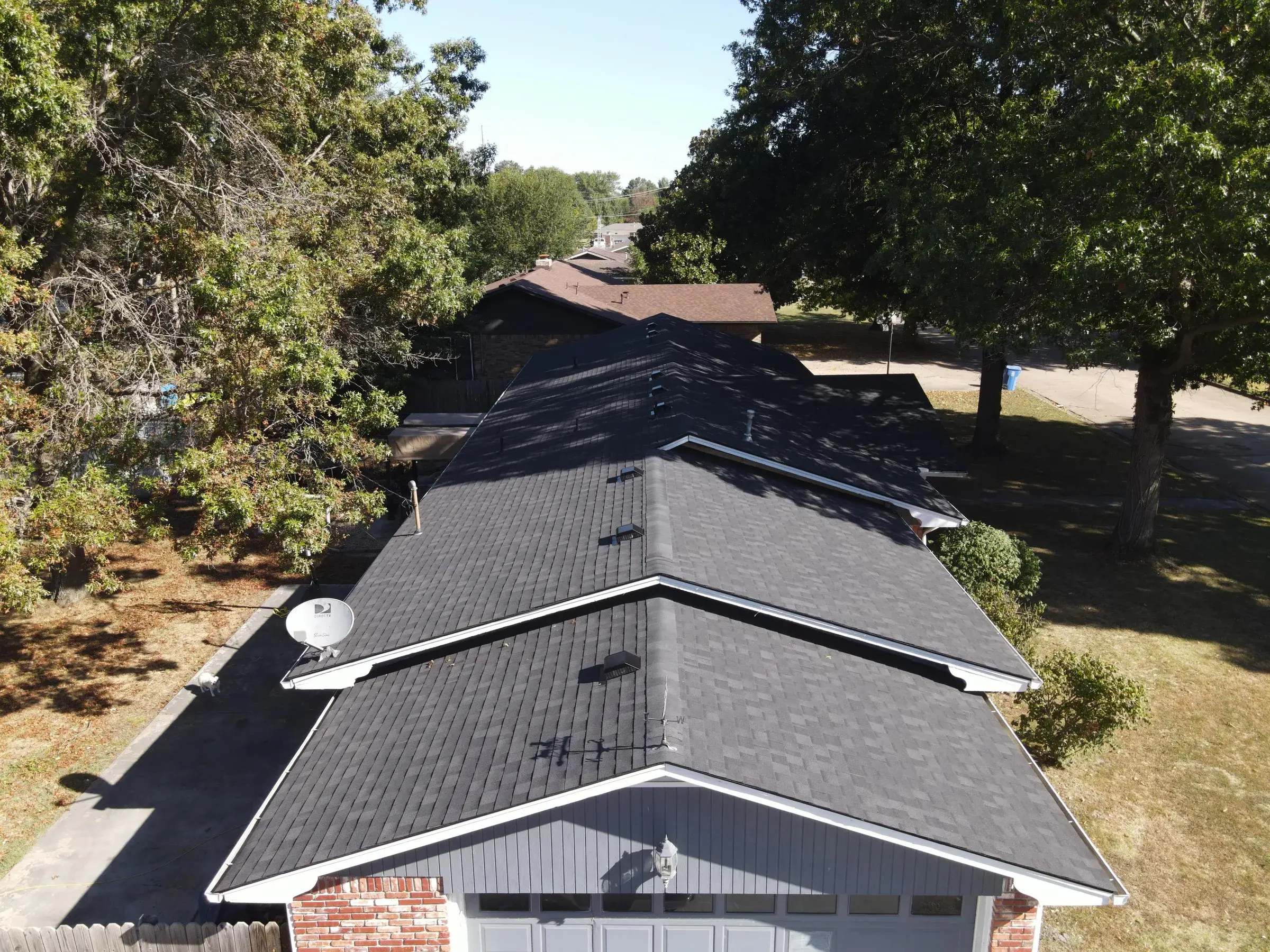 A drone view along the ridge of a house with a newly installed black asphalt shingle roof and white gutters, with a neighborhood street visible beyond.