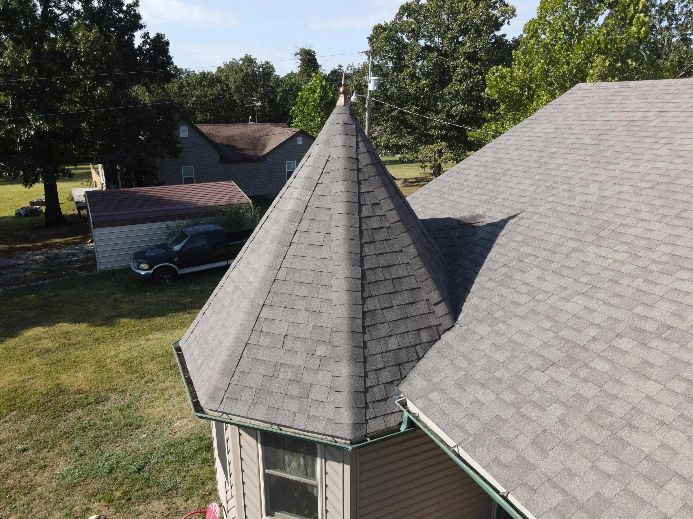 A drone view of a conical turret roof finished in gray asphalt shingles, meeting the larger shingled hip roof of a tan-sided house.