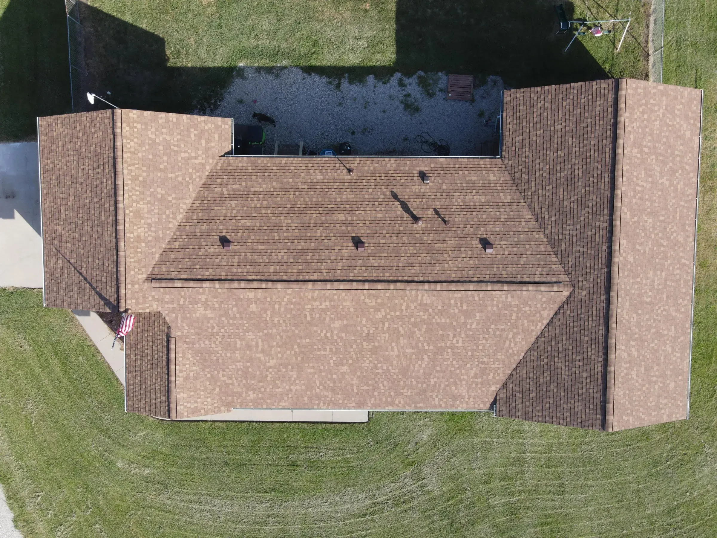 Overhead drone view of a home's completed tan asphalt shingle roof, showing clean hip lines and valleys surrounded by green lawn.