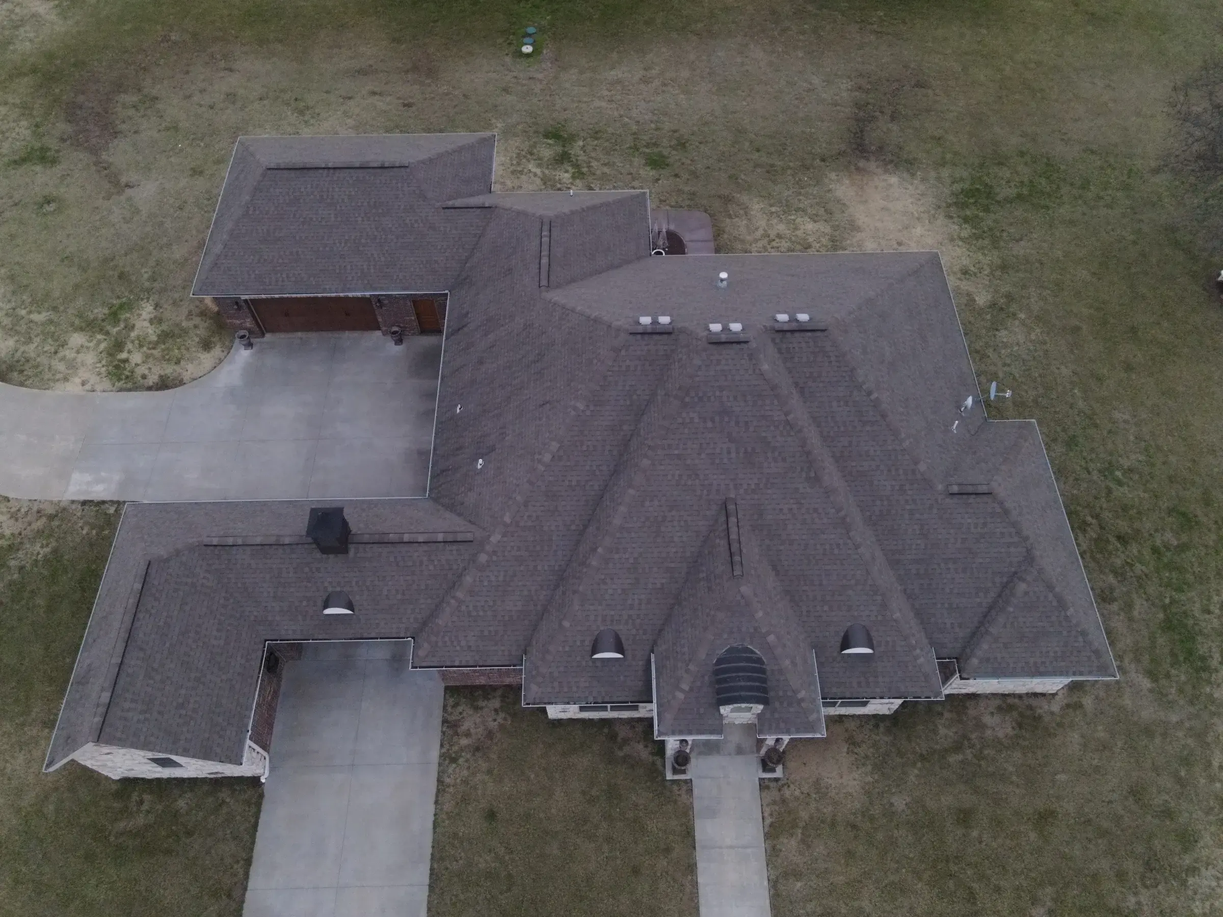 Aerial view of a large home with a complex multi-gabled roof covered in brown-gray architectural shingles, surrounded by dormant winter lawn.