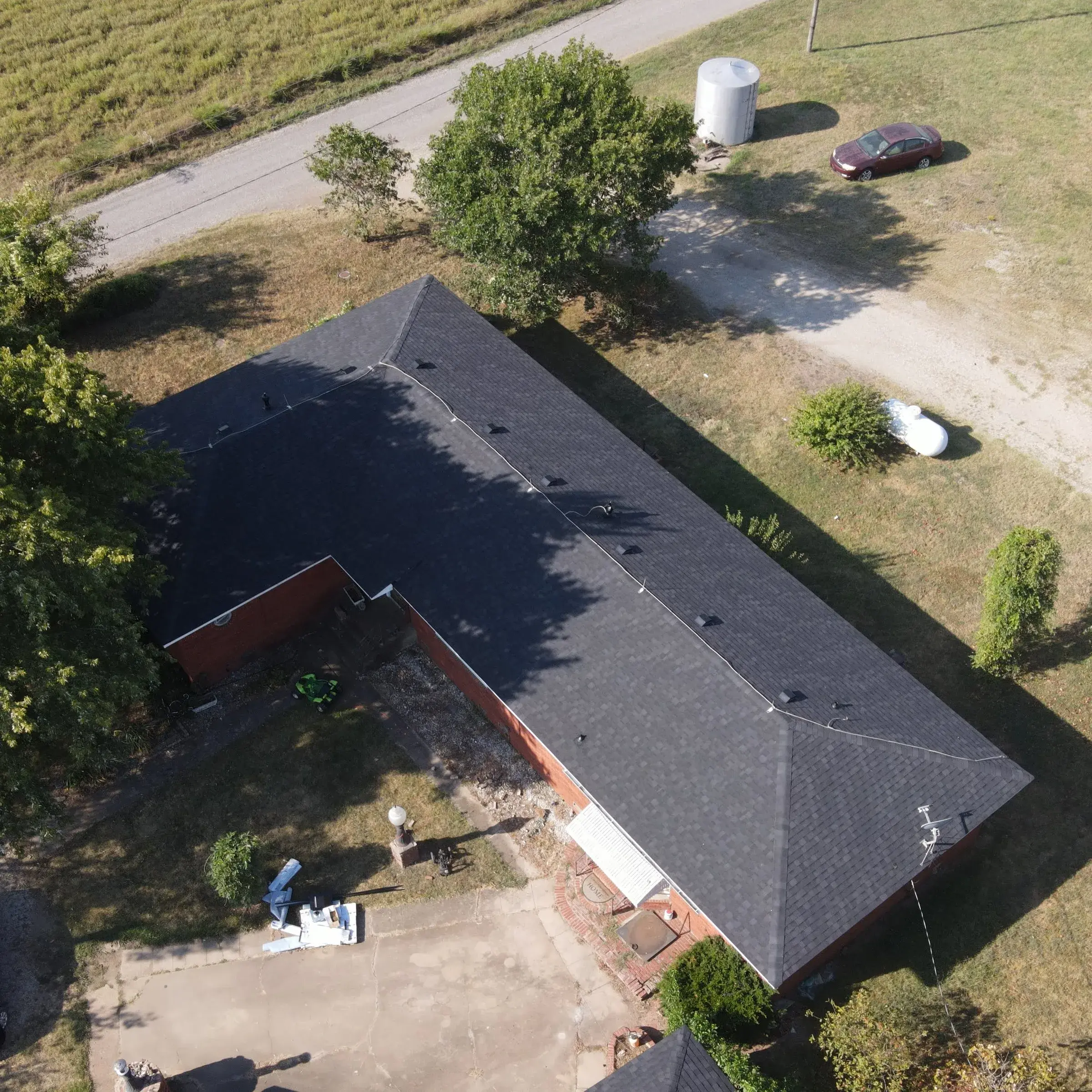 Aerial view of a long brick ranch home with a dark, uniform asphalt shingle roof, set on rural acreage with a gravel drive and storage tanks.