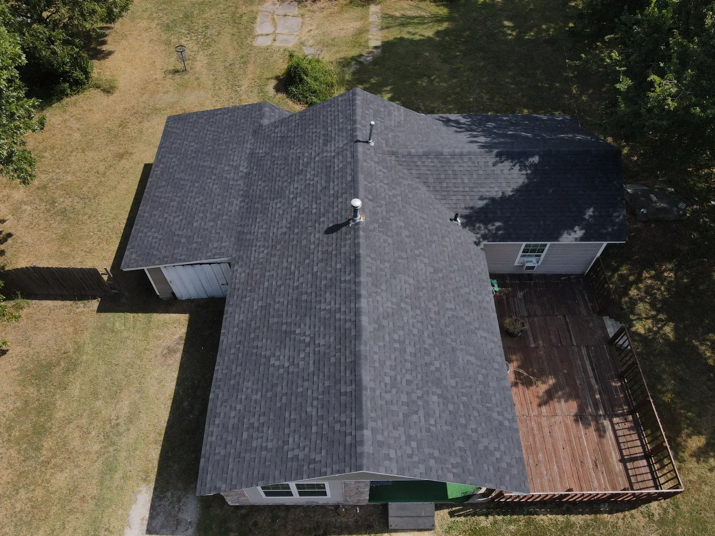 Overhead drone view of a ranch house with a clean dark gray asphalt shingle roof, a wood deck along one side and a metal carport at the other end.