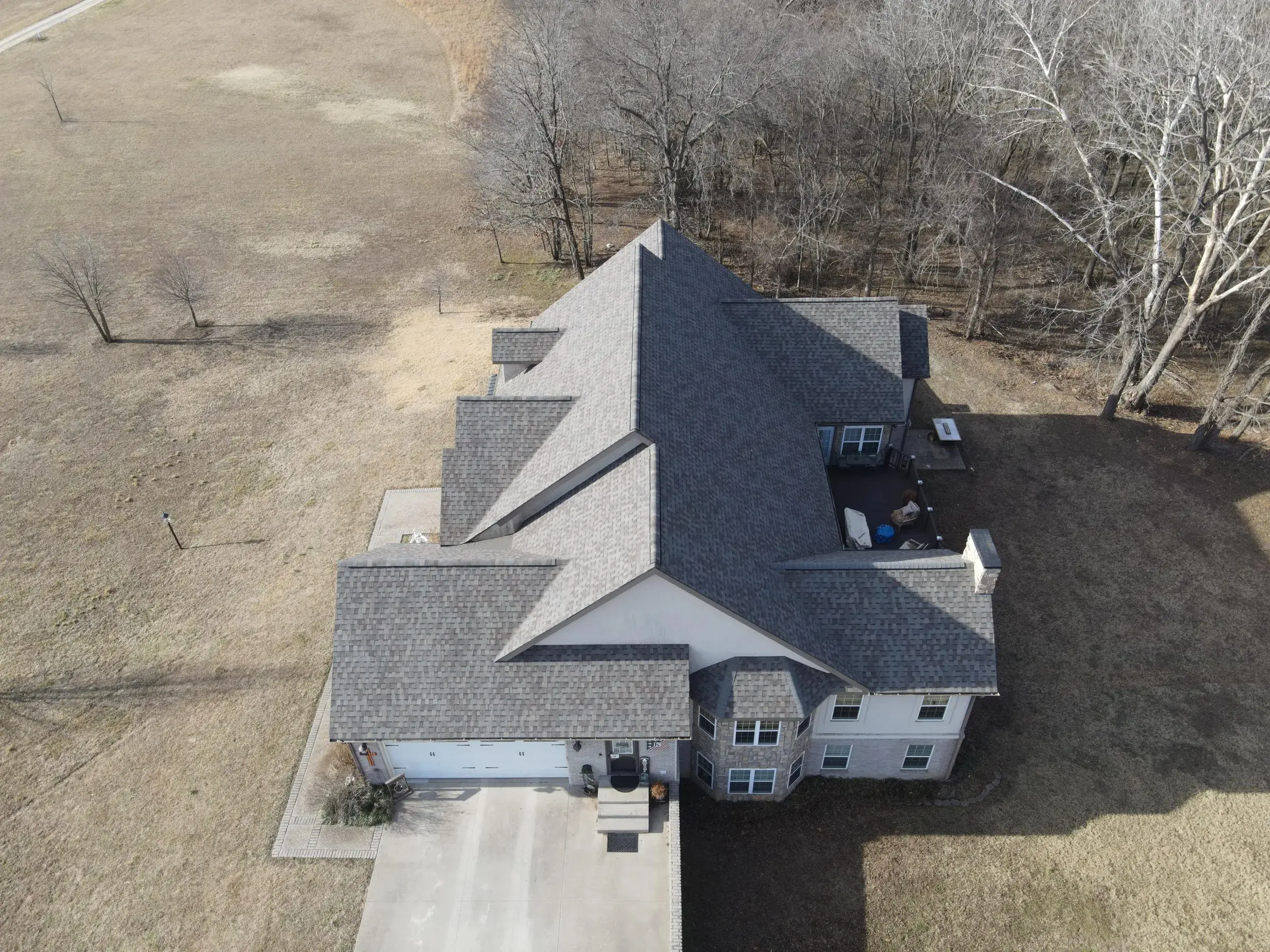 Aerial view of a large two-story house with a multi-gable gray architectural shingle roof, taken in winter with bare trees and dormant grass around it.
