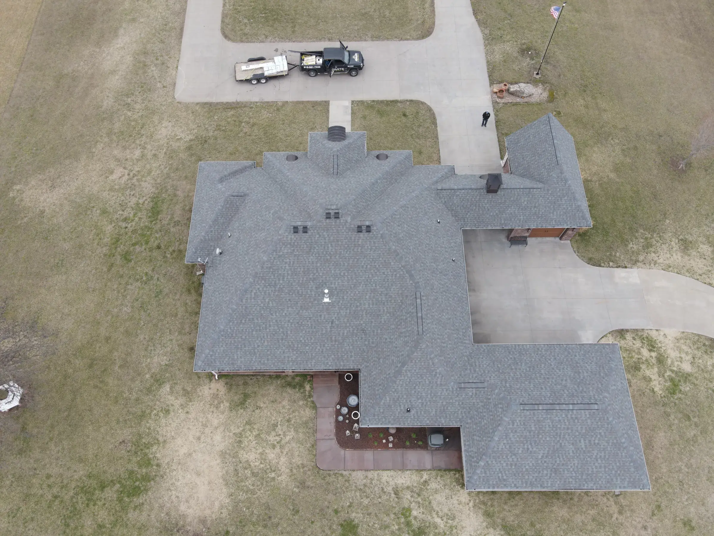 Aerial view of a large ranch home with a gray asphalt shingle roof, with a pickup truck and loaded dump trailer parked on the wide concrete driveway.