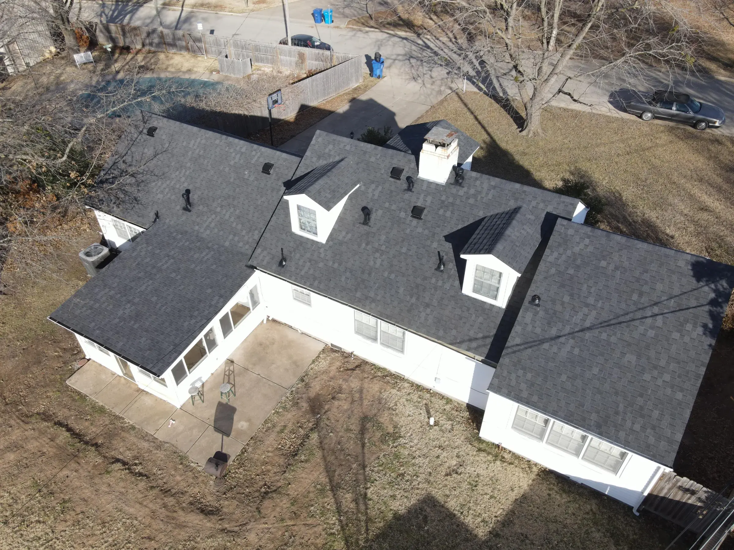 Aerial view of a white house with dormers and a dark gray asphalt shingle roof, with a fenced backyard and a street running behind it.