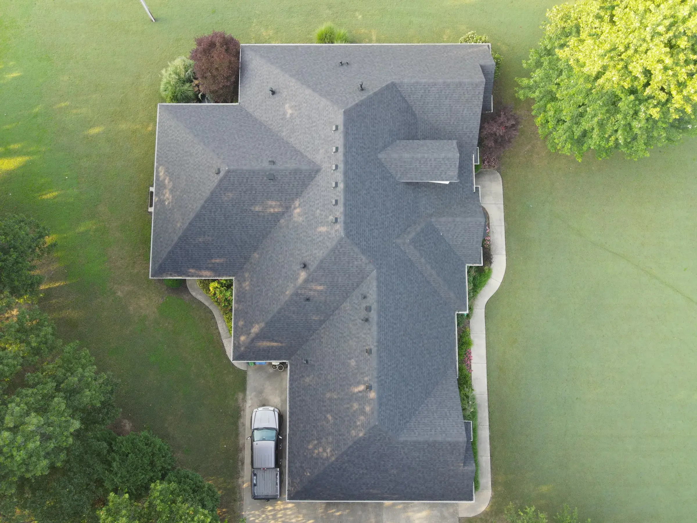 Overhead drone view of a large home with a clean dark charcoal asphalt shingle roof, surrounded by a manicured green lawn and mature trees.