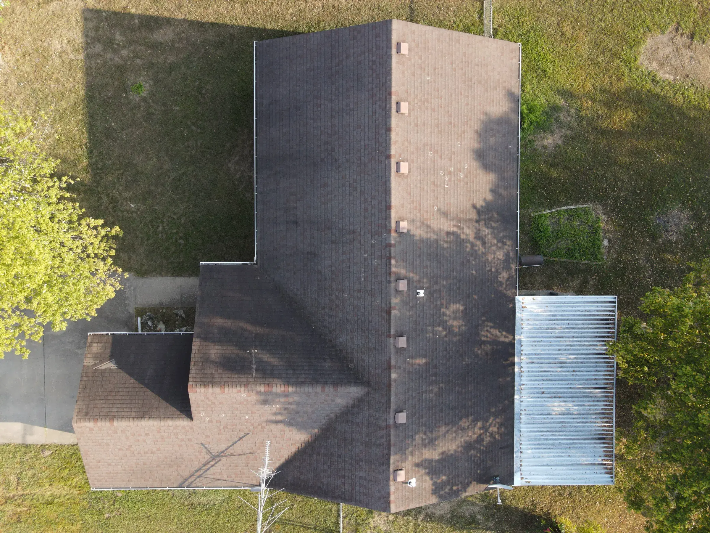 Overhead drone view of a large aging brown shingle roof with a row of vents along the ridge, next to a building with a corrugated metal roof.