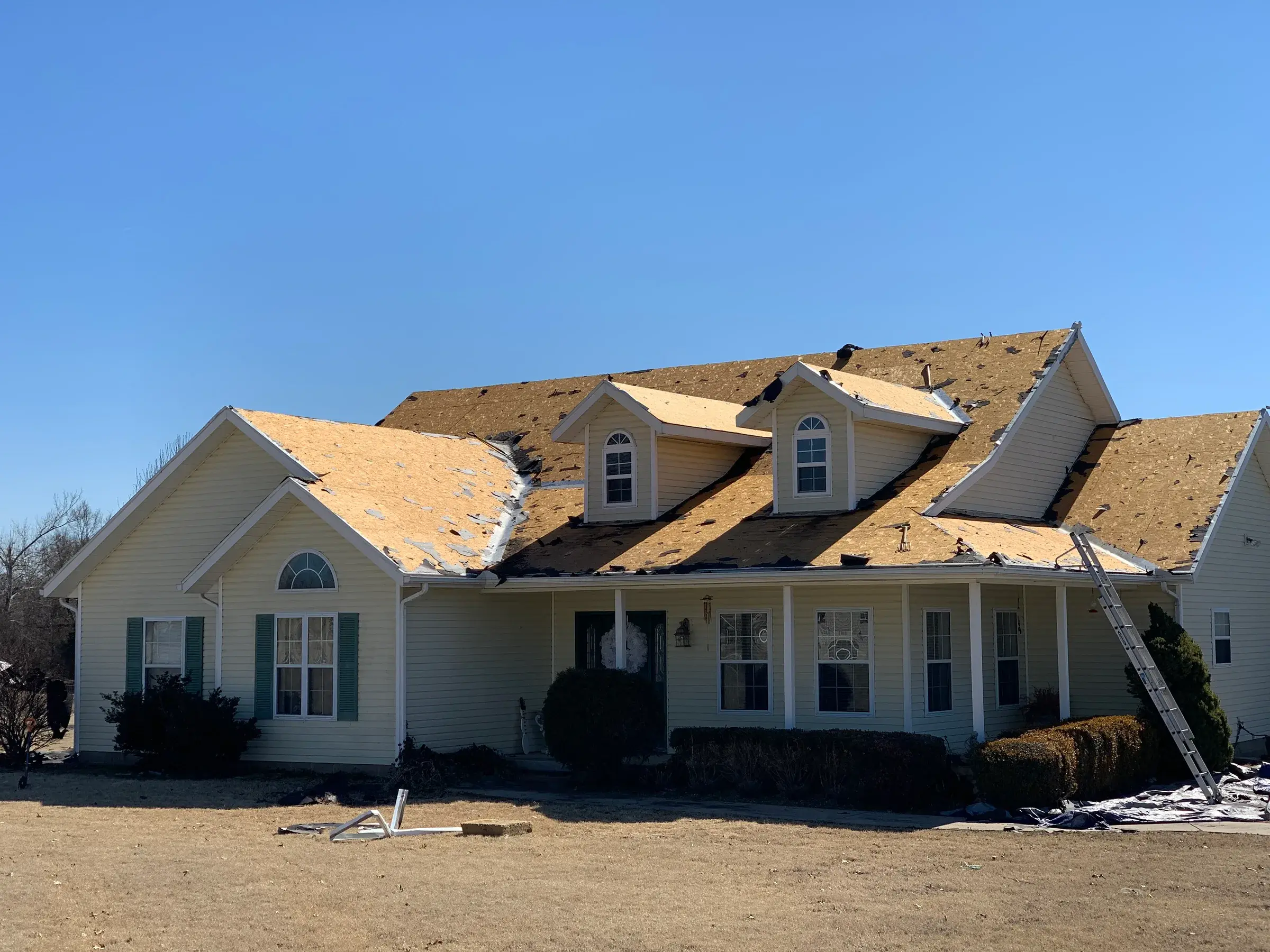 A large two-story house with its shingles stripped down to bare wood decking, with an extension ladder leaning against the eave.