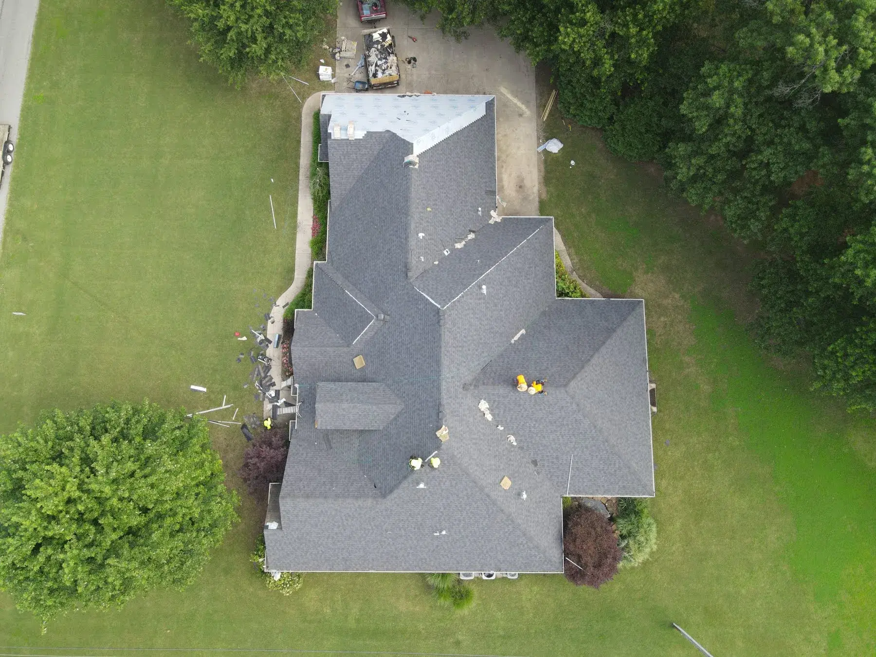 Aerial view of a large home mid roof replacement, with most planes covered in new dark charcoal shingles and one wing still showing white underlayment.