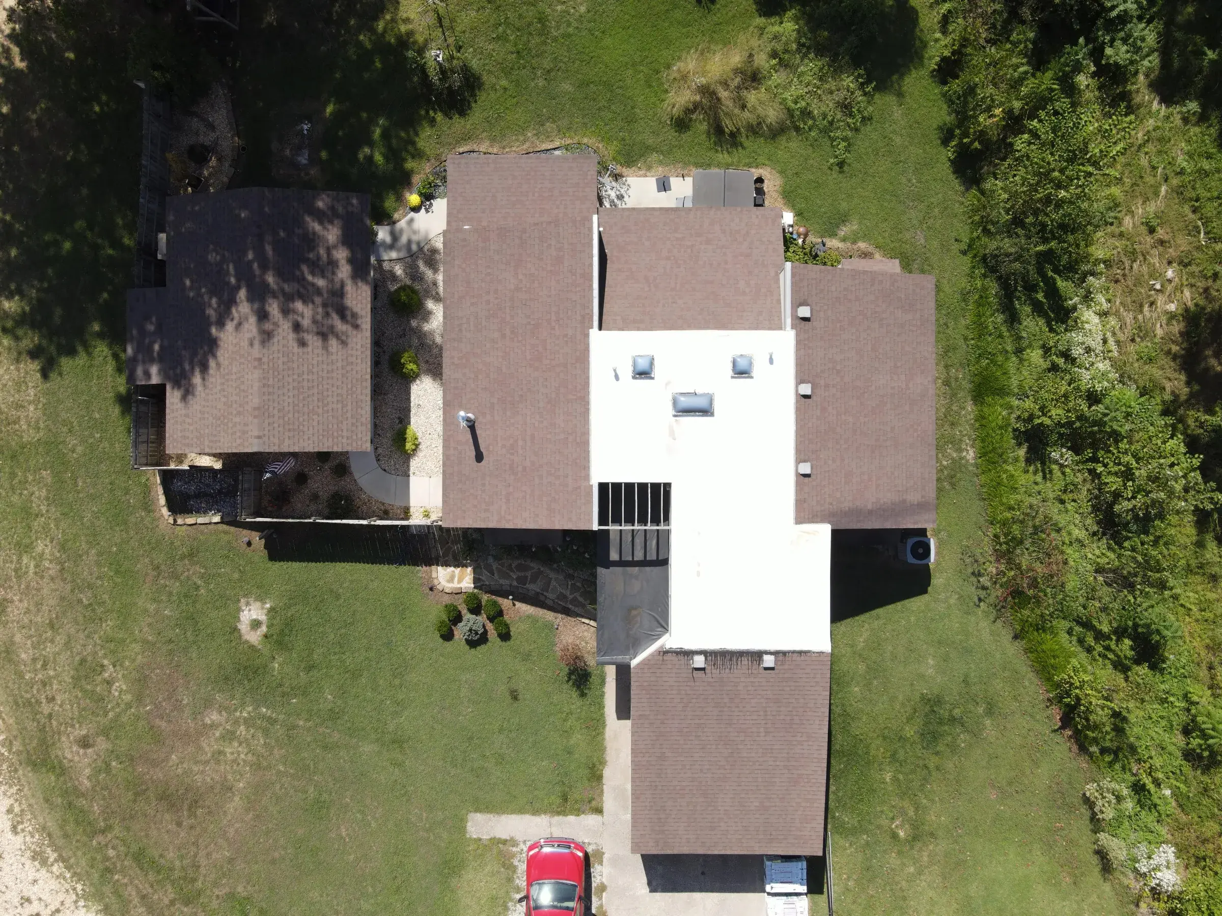 Overhead drone view of a house with a brown shingle roof wrapping around a white low-slope section fitted with several skylights.
