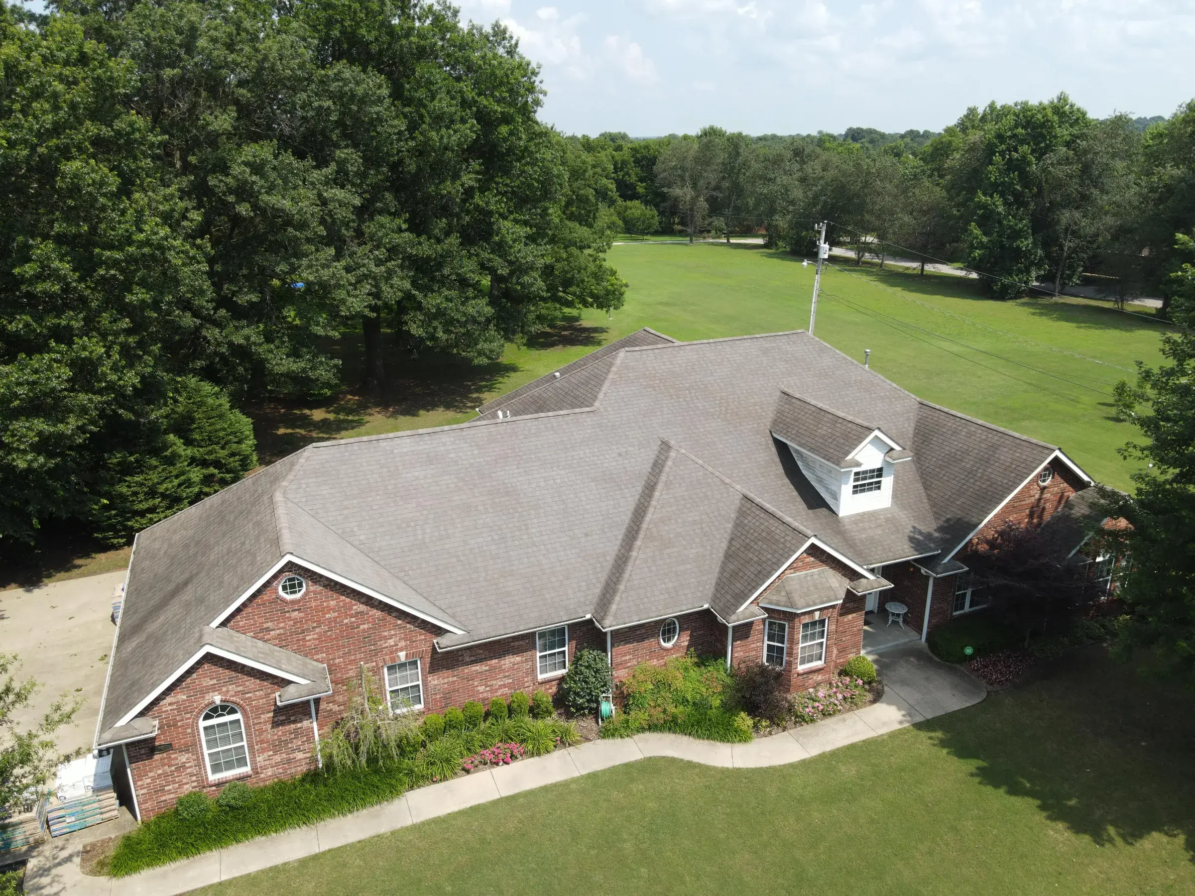 Aerial view of a large brick ranch home with a weathered gray-brown asphalt shingle roof, surrounded by mature oak trees and open field.