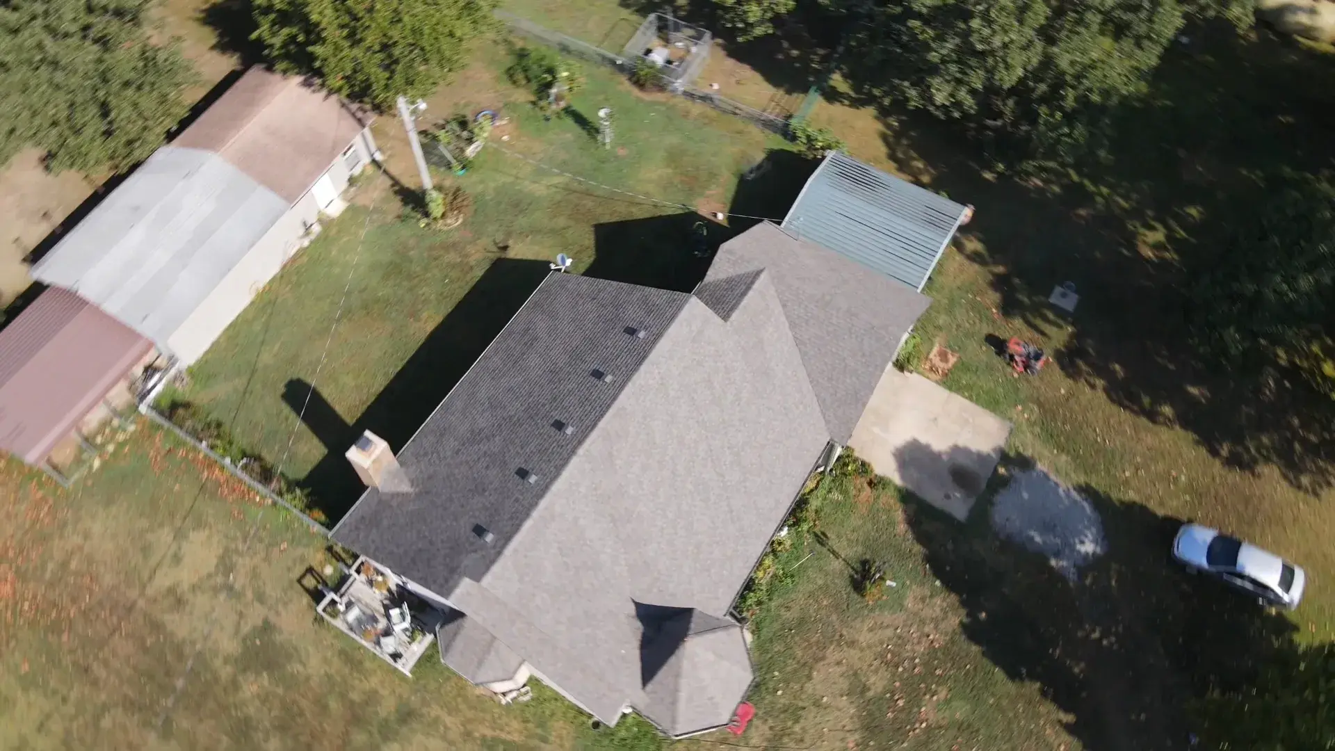 Overhead aerial view of a single-story rural home with a light gray shingle roof, outbuildings, and open grass around it.
