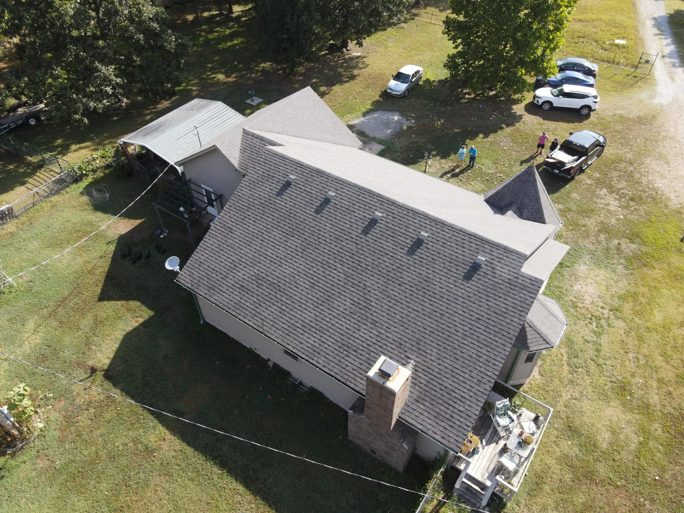 Angled drone view of a rural home with a light gray shingle roof, an adjoining metal carport, and vehicles parked on a gravel drive.
