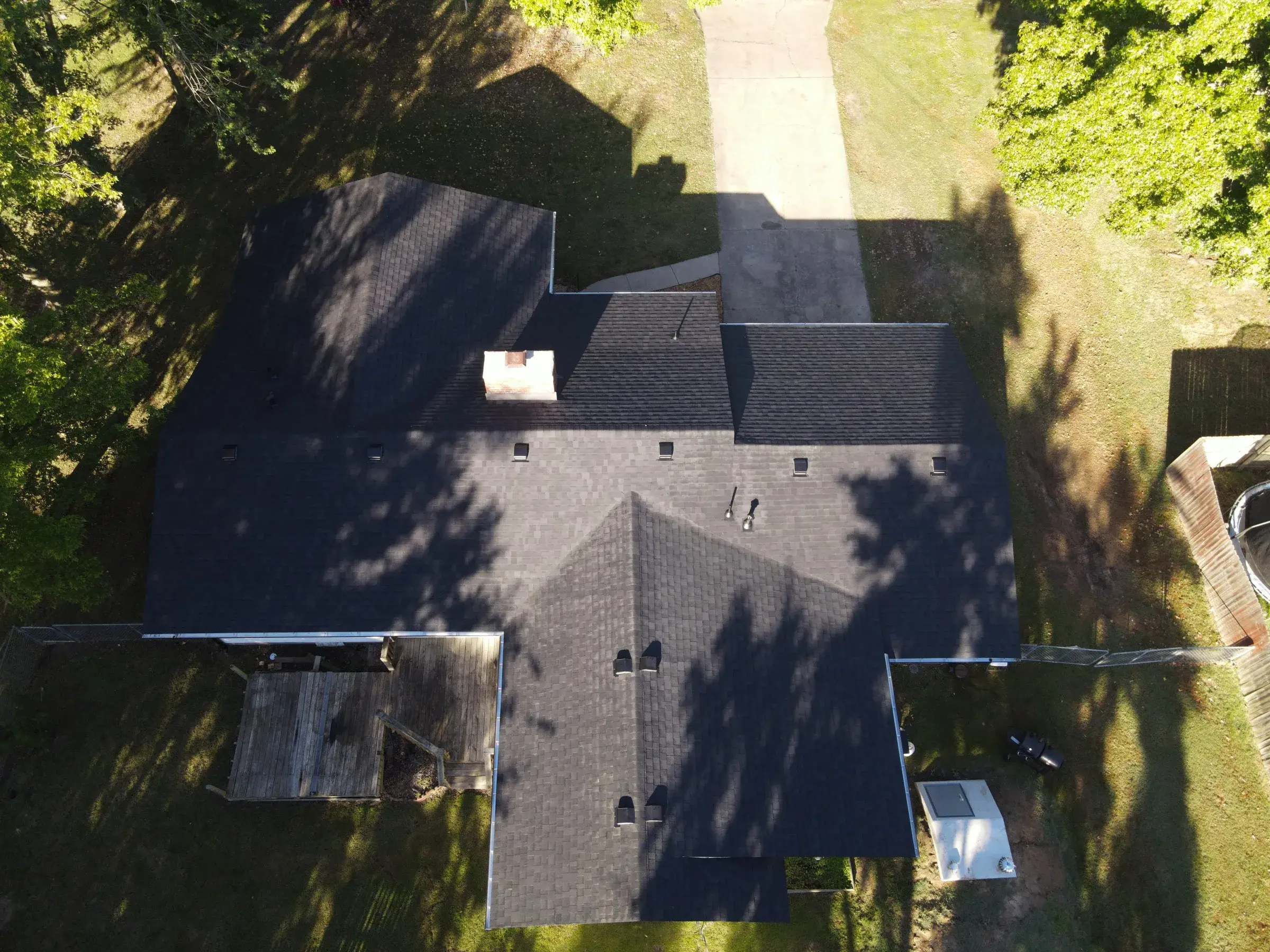 Overhead drone view of a house with a dark charcoal shingle roof across several gables, a brick chimney, and a wooden deck at one corner.