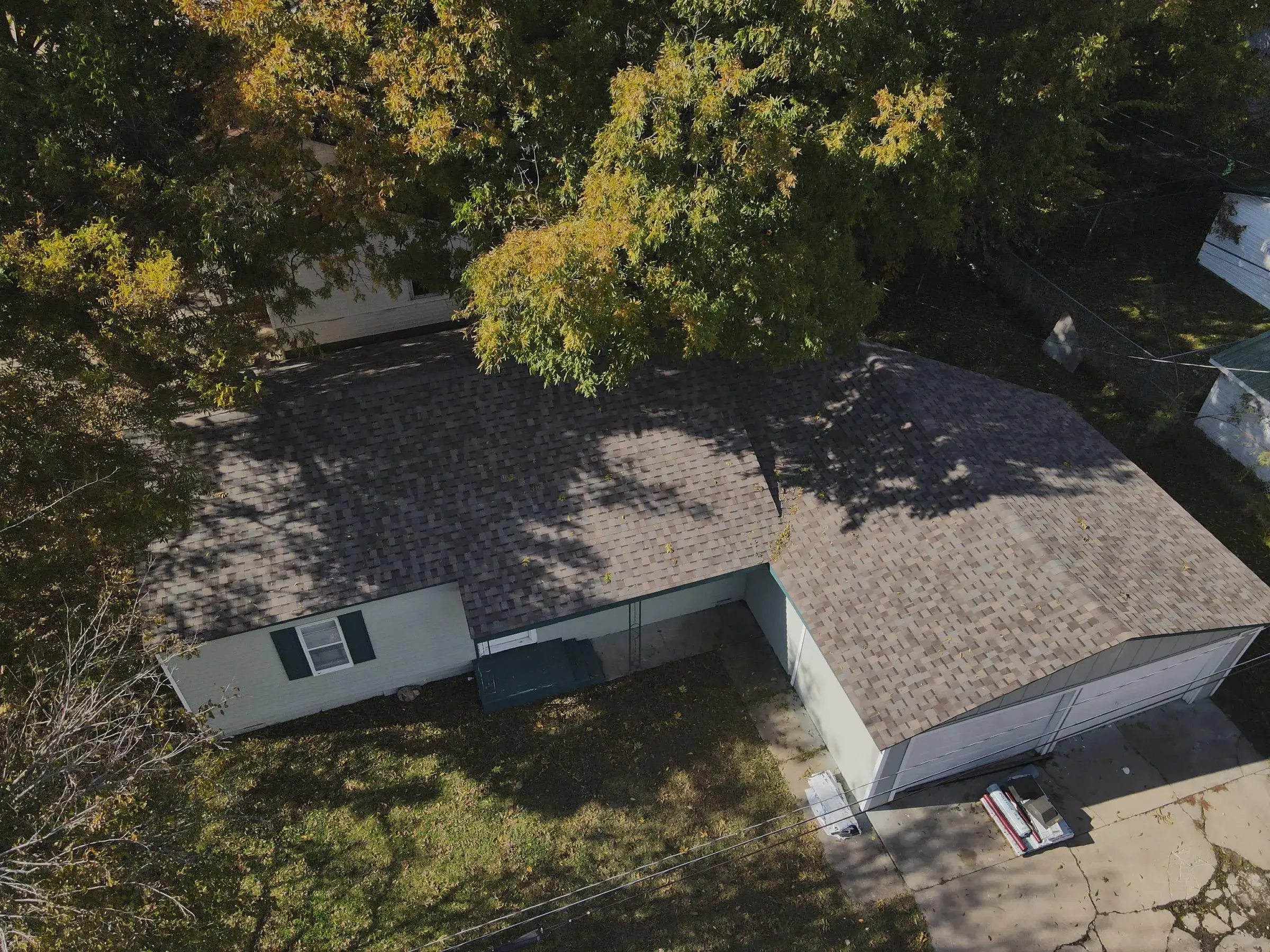 Angled drone view of a single-story home with a brown asphalt shingle roof and attached carport, partly shaded by a large tree in autumn.