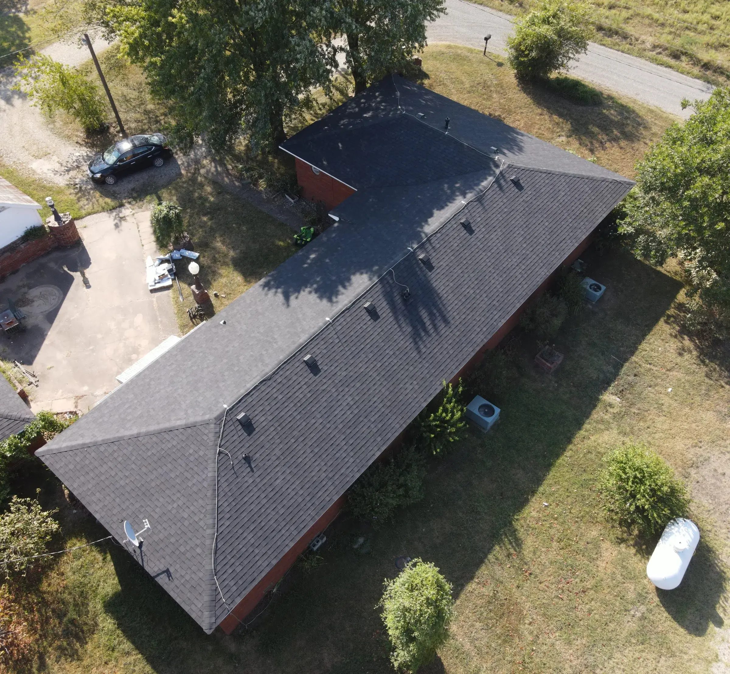 Aerial view of a long low brick building with a gray shingle roof and a row of ridge vents, with a car parked on the gravel drive alongside.