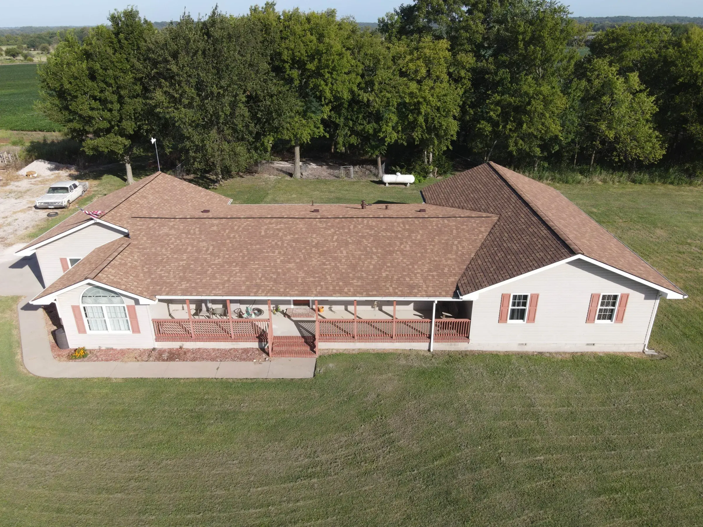 Aerial view of a cream ranch-style home with a brown shingle hip roof and a long covered front porch with red railings, set on a green lawn with farmland behind.