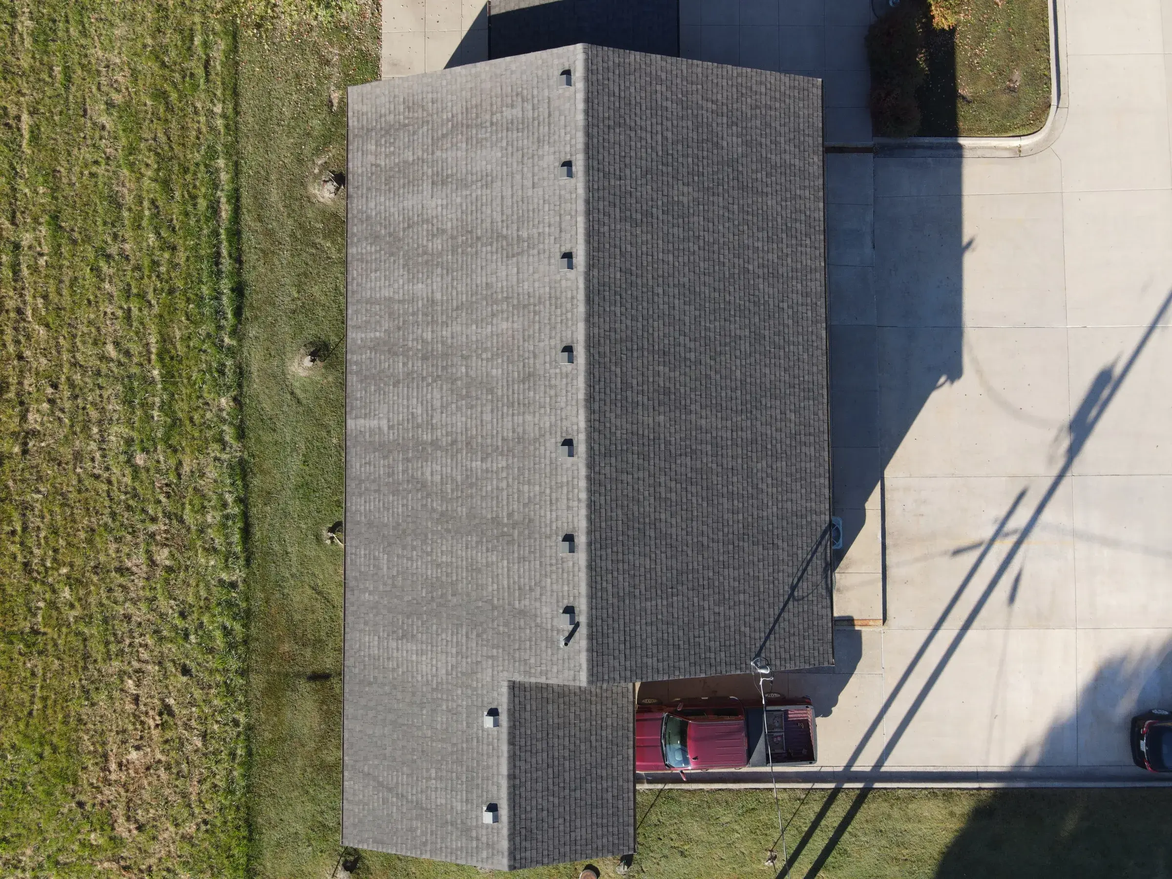 Overhead view of a long narrow building with a light gray shingled gable roof beside a large concrete drive.