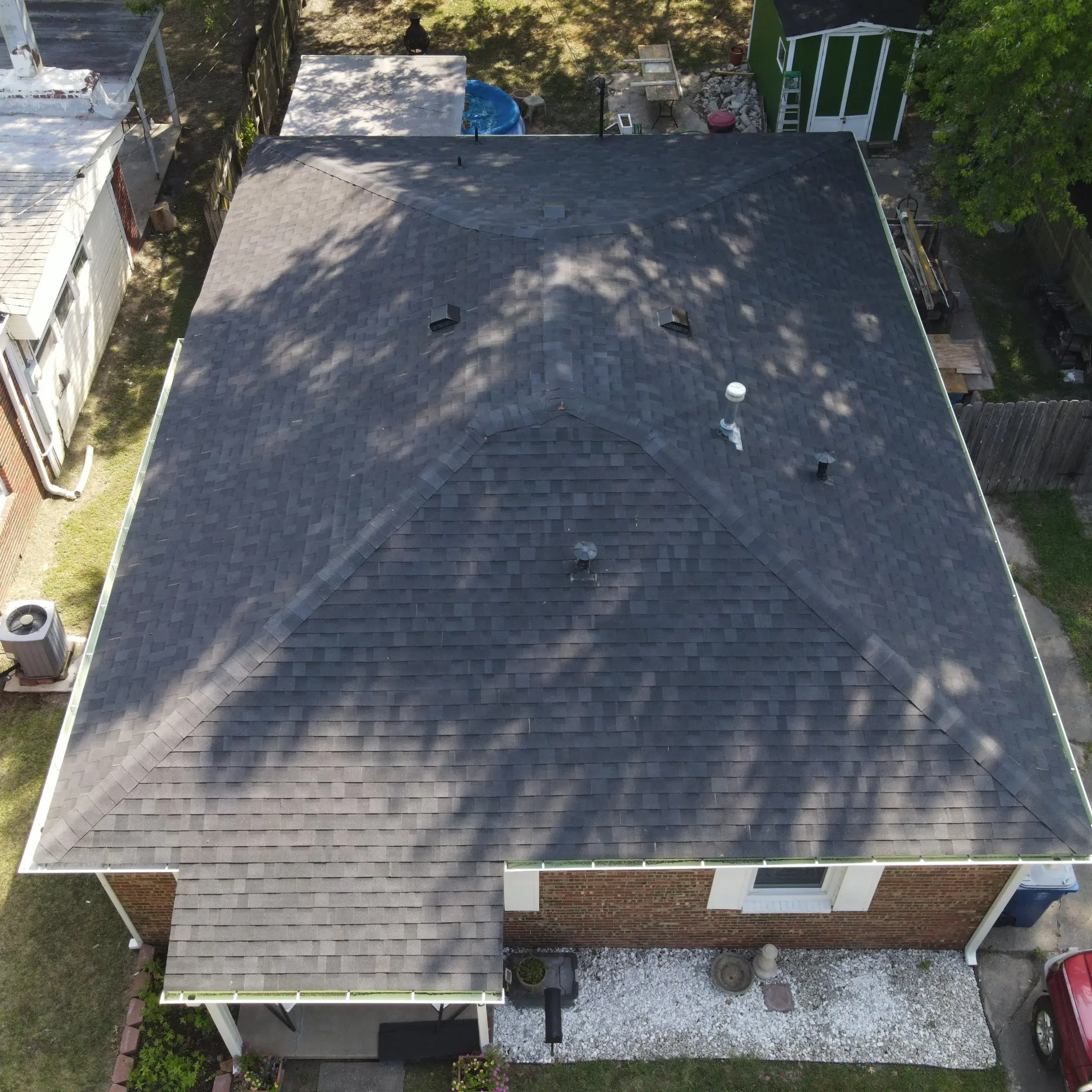 Overhead drone view of a brick house with a dark gray shingle hip roof, dappled with tree shadows, with a shed and neighboring yards visible around it.