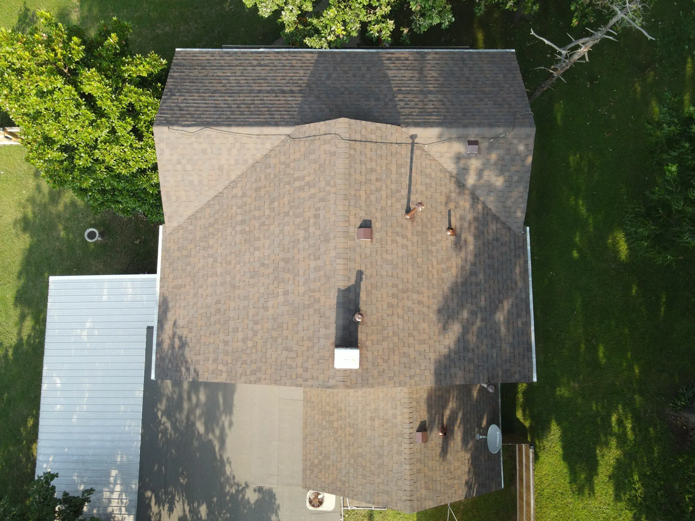 Overhead drone view of a house with a tan and brown architectural shingle roof, with pipe vents and a flue on the slope and a metal-roofed outbuilding at the corner of the frame.