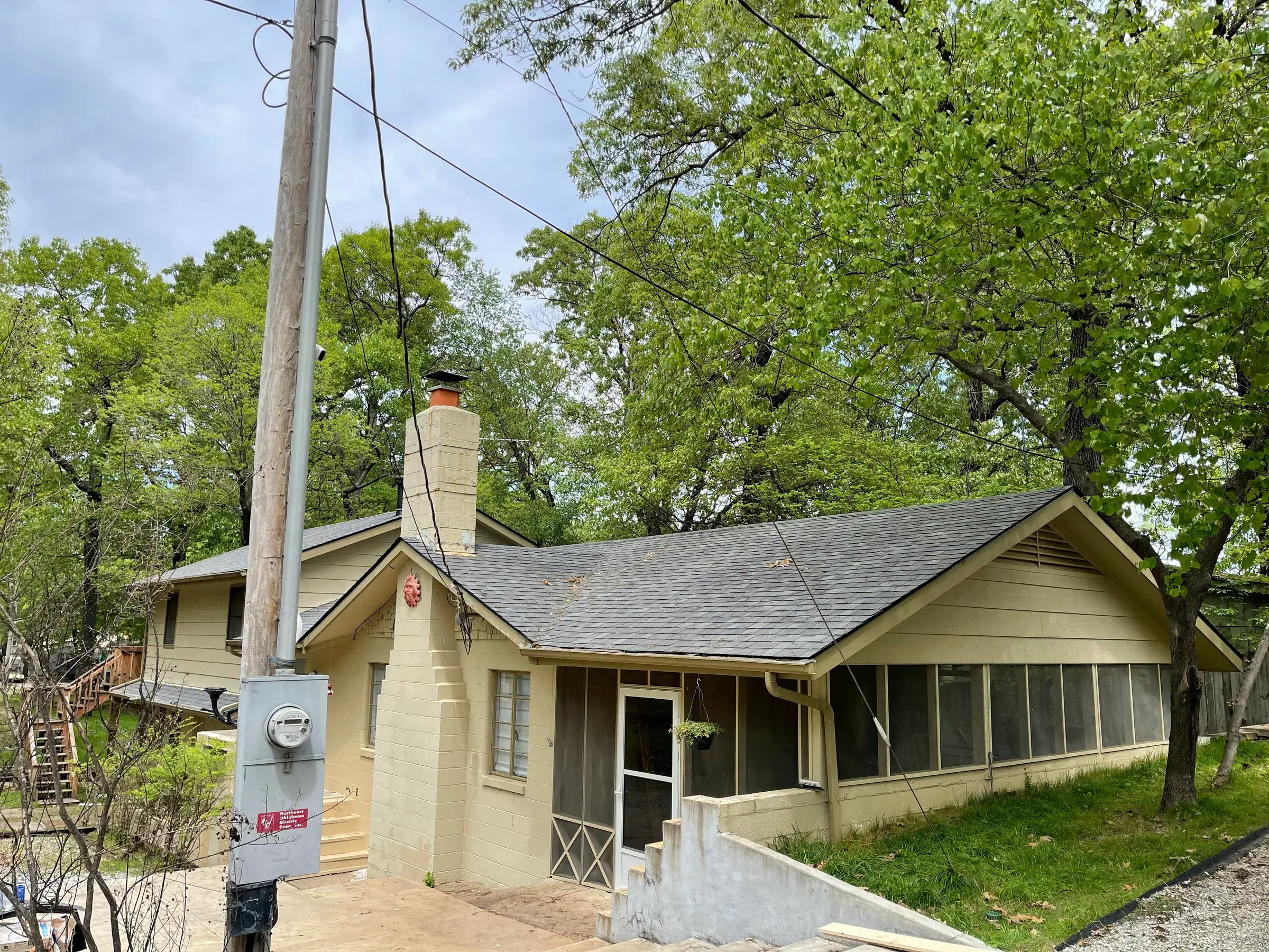 Single-story olive-green house with a new dark asphalt shingle roof and stone chimney, framed by a utility pole and overhead power lines in the foreground.