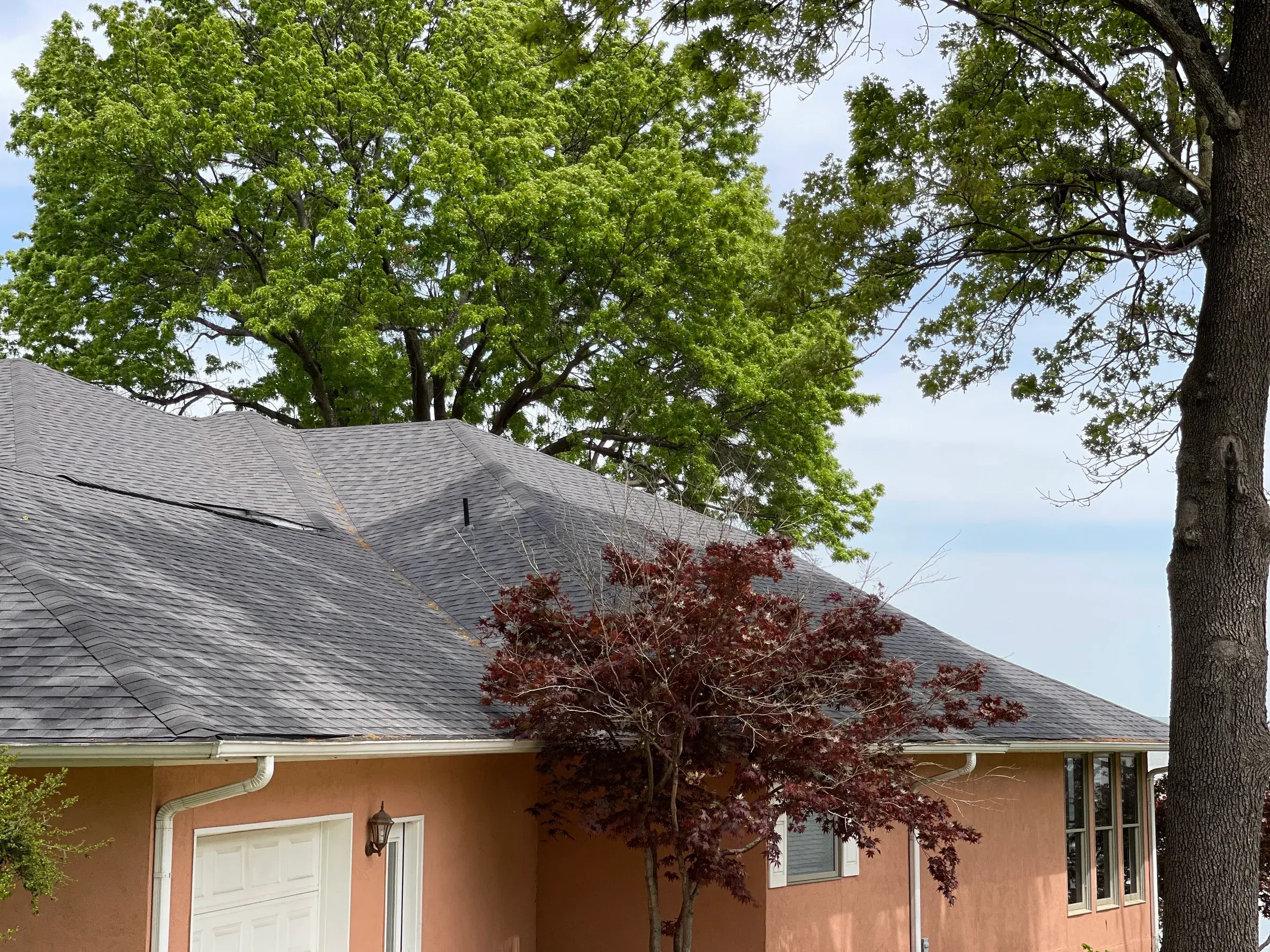 Aged asphalt shingle hip roof on a peach stucco home, with mature trees overhanging the ridge and uneven patches across the shingle field.